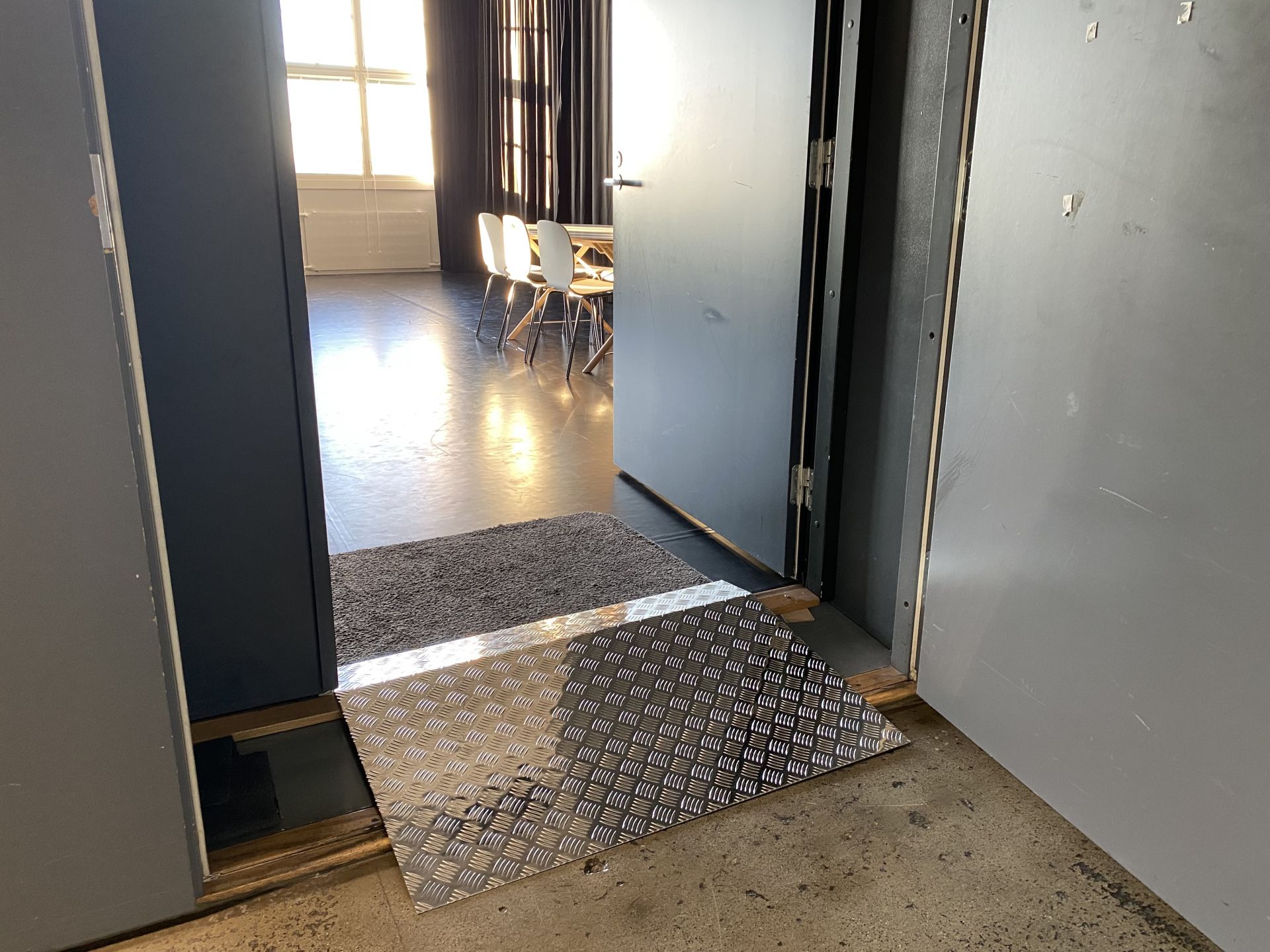 En dörröppning med en rullstolsramp i metall. I rummet lyser solen in genom fönster som skymtar.