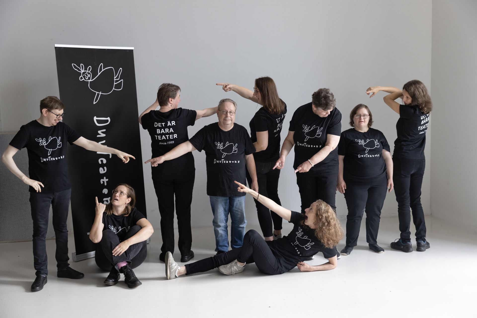 Bilden visar en grupp på åtta personer som poserar framför en bakgrund med en svart banner där det står "DuvTeatern”. De har alla på sig svarta t-shirts med en enkel illustration av en duva och texten "DuvTeatern”. Några personer står med ryggen mot kameran och då syns texten på T-skjortans rygg ”Det är bara teater” Personerna står i olika positioner och pekar mot varandra i en lekfull och dynamisk gruppbild. Några av personerna sitter på golvet, medan andra står och pekar eller rör sig på olika sätt. 