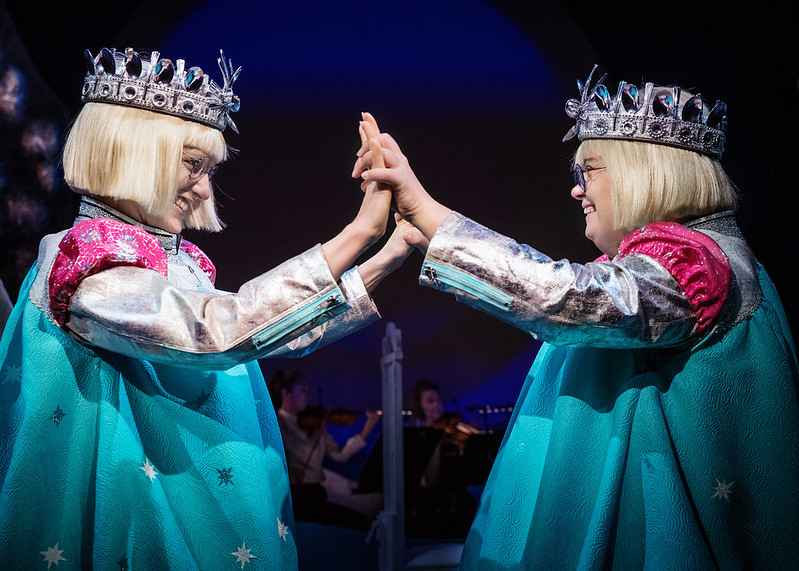 Actresses Sophia Heikkilä and Karolina Karanen in their princess roles. The princesses stand facing each other, holding hands against each other. They wear crowns, blonde bob haircuts, and beautiful silver, pink, and turquoise-colored dresses.
