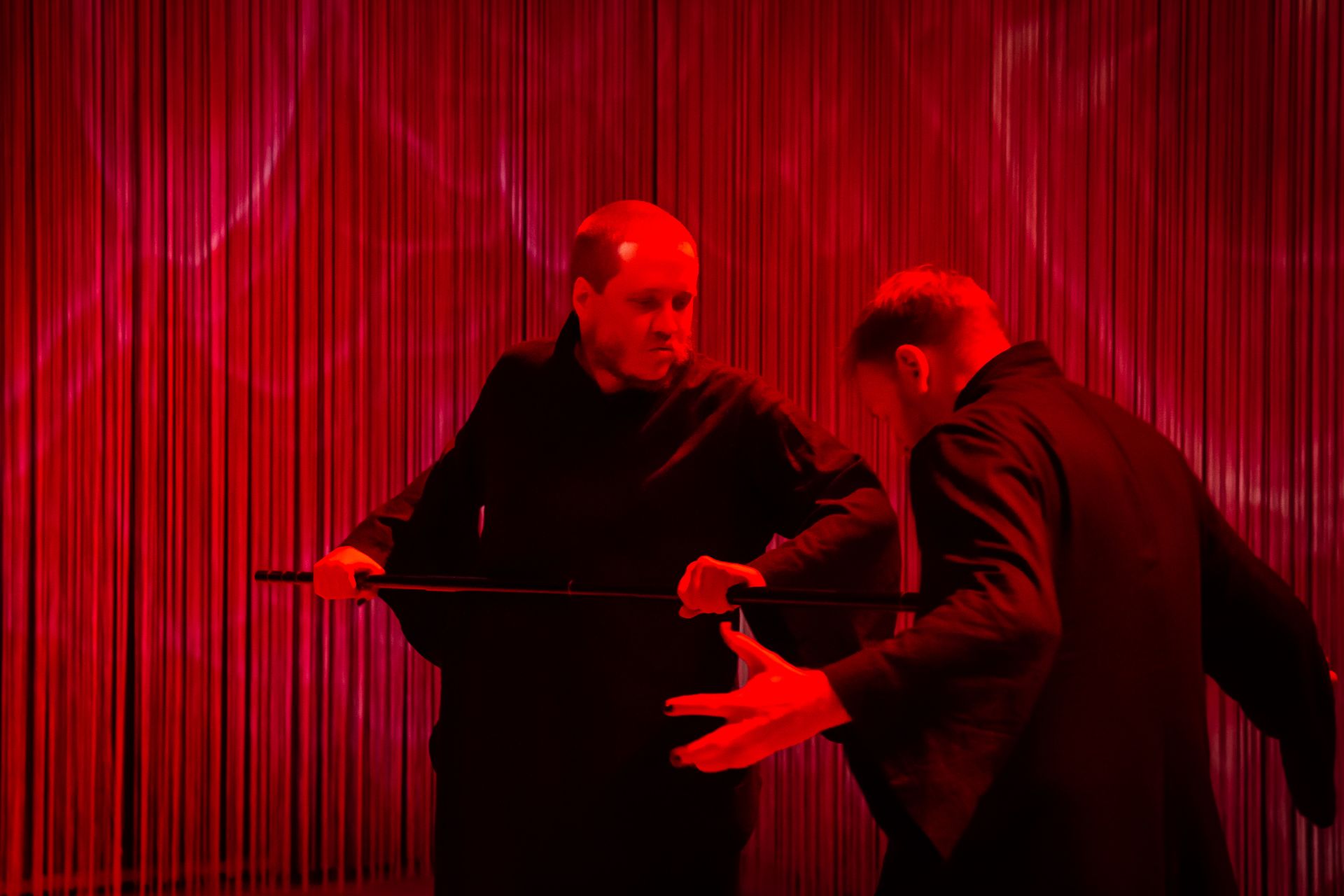 Föreställningsbild där dansare Emil sticker en käpp mot dansare Eero. Båda dansarna är svartklädda och scenen är inbäddad i ett dramatiskt mörkrött ljus.